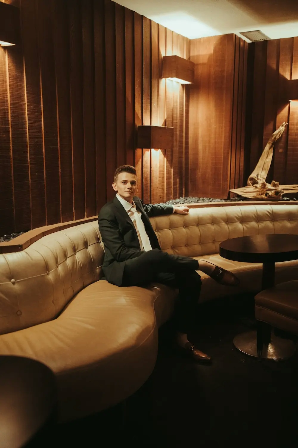A picture of John, seated on a couch in a lounge wearing a dark green suit.